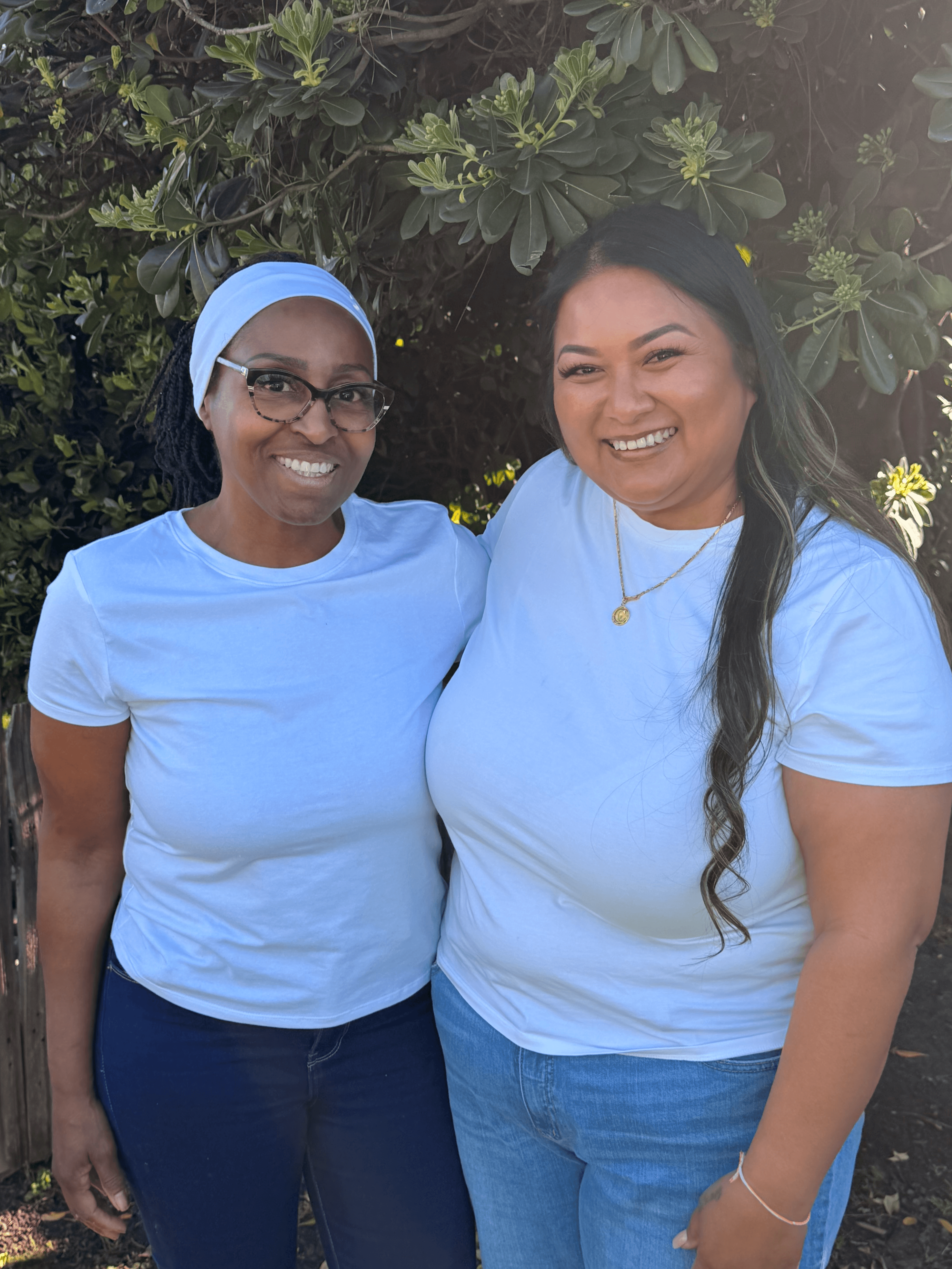 Two women wearing white t-shirts smile while standing outdoors in front of green bushes.